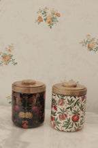 Two vintage ceramic canisters with wooden lids, one featuring a dark floral pattern and the other a white pomegranate and leaf print, displayed on a floral wallpaper background.